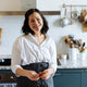 Emiko Davies is standing in a kitchen smiling. There are cooking utensils on the wall and benchtop behind her
